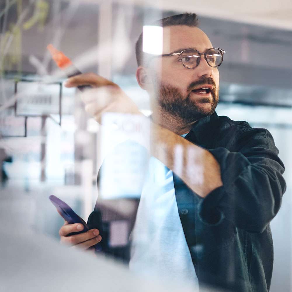 Blick durch eine Glasscheibe auf einen beschäftigten männlichen Unternehmer mit Brille, der auf eine transparente Tafel zeichnet, während er seine Geschäftsstrategie während einer Besprechung im Büro erläutert.
